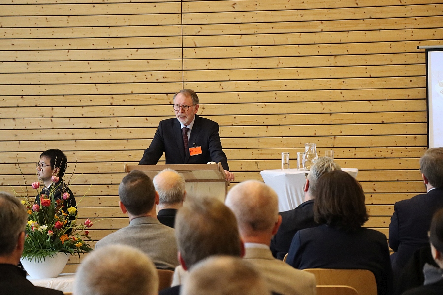 Das Bild zeigt einen Mann am Rednerpult. Er spricht zu den Gästen, die auf Stühlen vor ihm sitzen. 