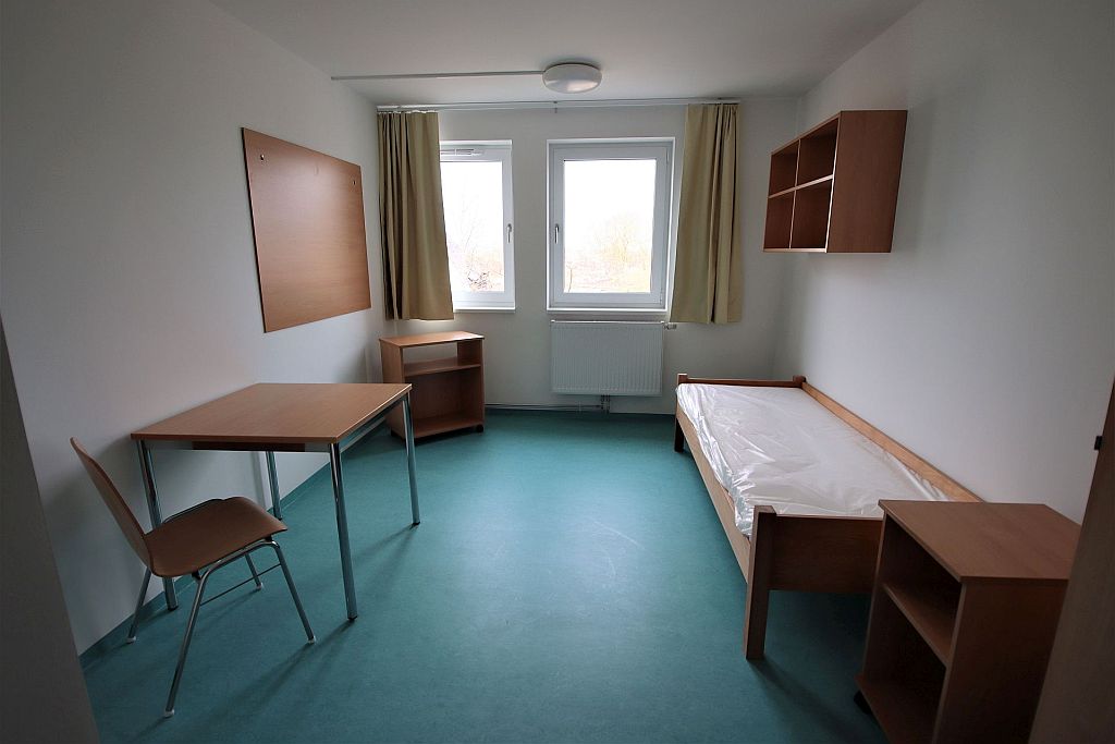 Das Bild zeigt ein eingerichtetes Einzelzimmer mit Bett, Tisch, Regal und Schrank.