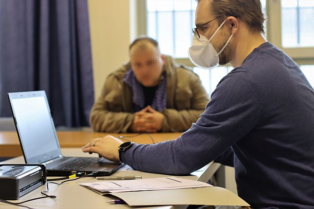 Ein Mann sitzt am Schreibtisch vor einem Laptop. Ein zweiter Mann sitzt neben ihm am Tisch und beantragt einen Personalausweis.