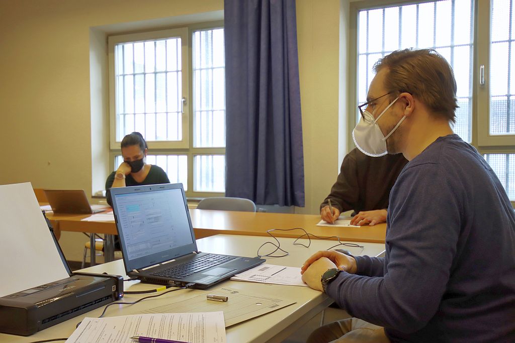 Ein Mann sitzt an einem Schreibtisch vor einem Laptop. Im Raum sind weiterhin eine Mitarbeiterin der JVA Stralsund und ein Gefangener. Der Gefangene beantragt einen neuen Personalausweis und unterschreibt ein Formular.