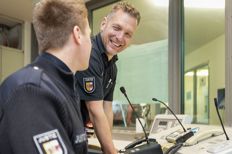 Zwei Justizvollzugsbeamte in Uniform unterhalten sich freundlich in einem Büro mit Gegensprechanlage. Einer der Beamten lehnt sich lächelnd über einen Schreibtisch mit Kommunikationsgeräten.